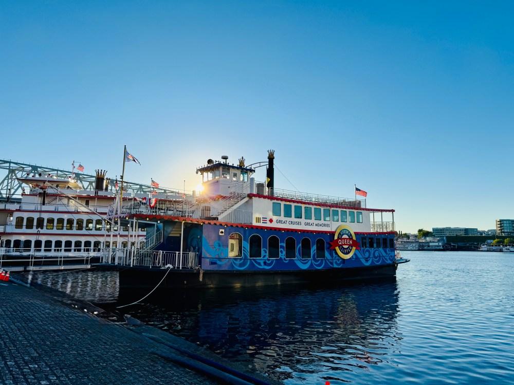 Colorful riverboat docked, flags flying, under a clear blue sky with sunlight behind.