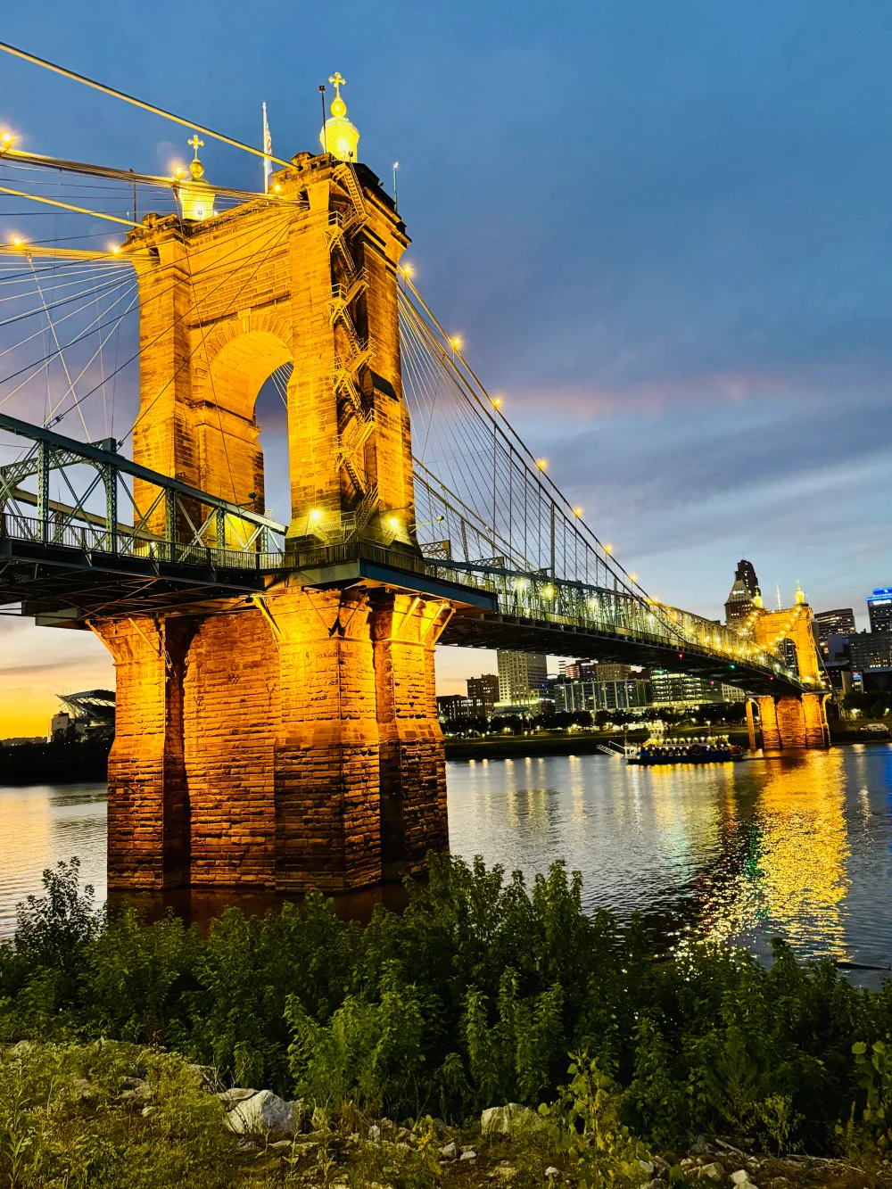 Illuminated suspension bridge over river at dusk with city skyline in the background.
