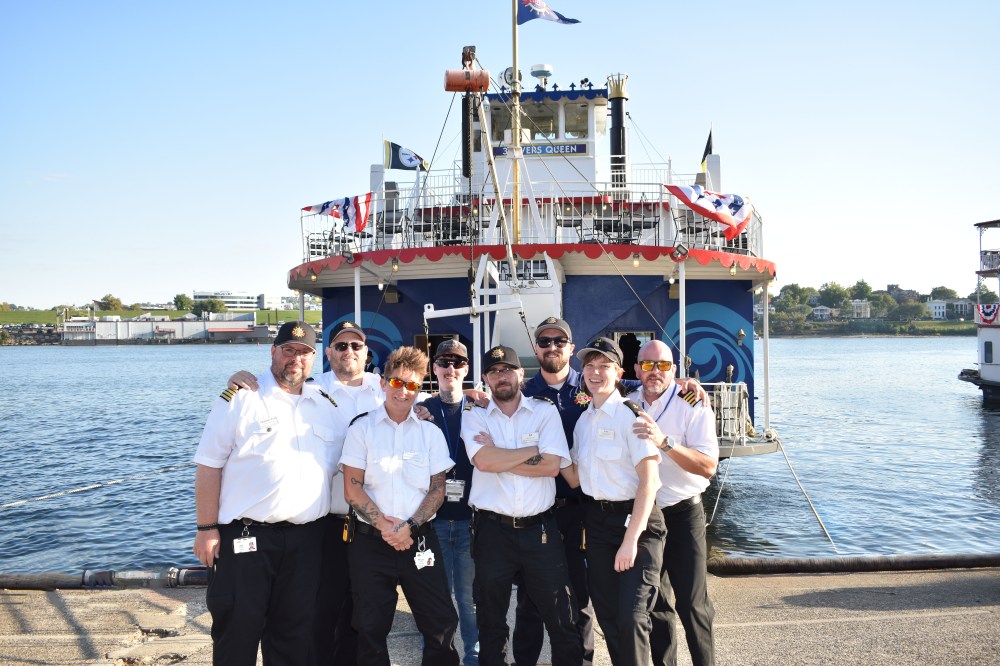 Eight crew members in uniforms pose in front of the Patriot Harbor Lines vessel at a dock.